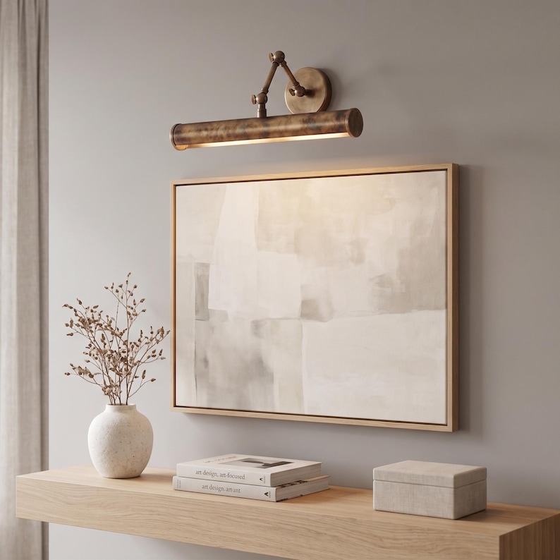 May include: A bronze-toned picture light is mounted above a framed abstract painting. A white vase with dried branches, two books, and a small box sit on a wooden shelf. The painting has a neutral color palette.