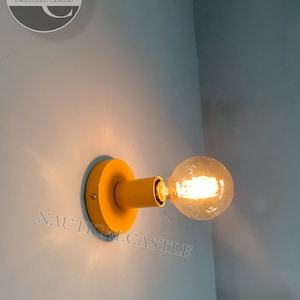 May include: A yellow wall sconce with a clear glass light bulb. The sconce is mounted on a wall and has a round base. The light bulb is turned on and is emitting a warm glow. The text "NAUTICALCASTLE" is visible in the lower right corner of the image.