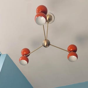 May include: A modern ceiling light fixture with a mid-century modern design. The fixture features six orange-red spherical shades with white interiors, connected by brass-colored arms to a central sphere. The light is mounted on a white ceiling.
