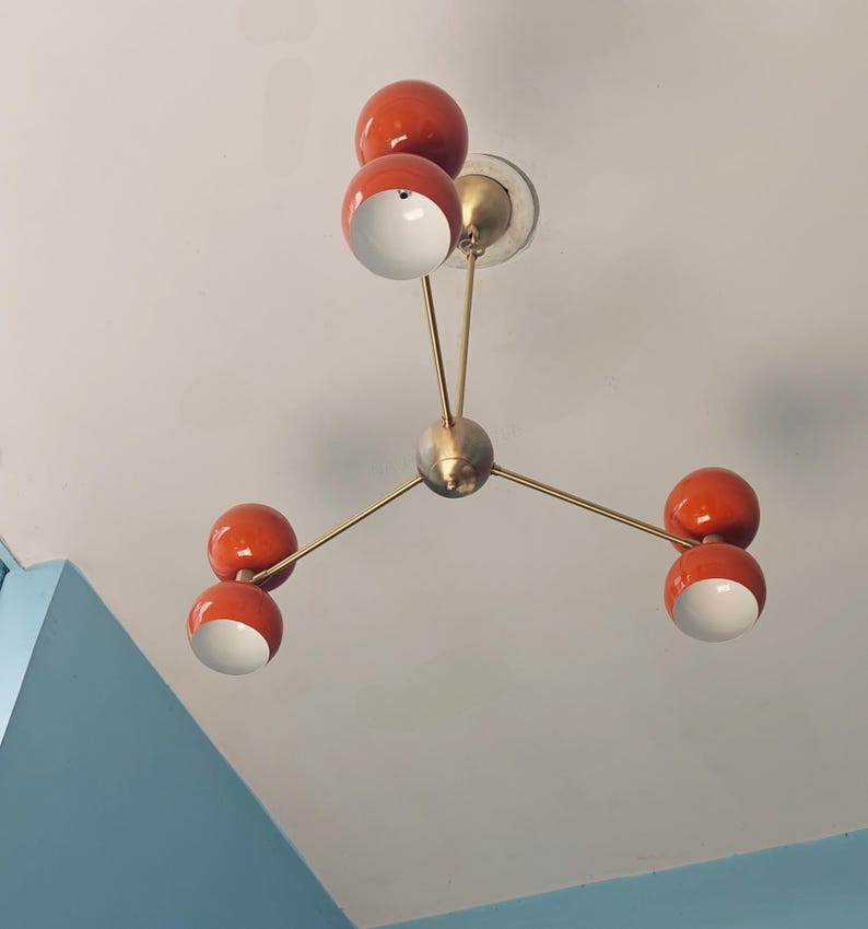 May include: A modern ceiling light fixture with a mid-century modern design. The fixture features six orange-red spherical shades with white interiors, connected by brass-colored arms to a central sphere. The light is mounted on a white ceiling.