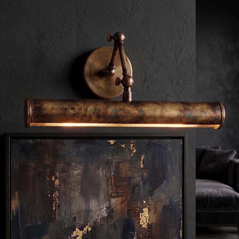 May include: A vintage-style wall-mounted picture light with a bronze finish. The light has a cylindrical shade and an adjustable arm, mounted on a circular backplate. The light is positioned above a framed painting.