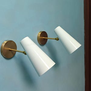 Peut inclure: Deux appliques murales blanches en forme de cône avec des plaques arrière et des bras circulaires de couleur laiton. Les appliques sont montées sur un mur bleu clair. Le texte "NAUTIC AST" est visible.