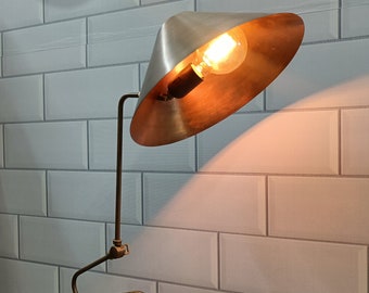 May include: A silver and brass desk lamp with a cone-shaped shade. The lamp is on a white marble base and is illuminated by a light bulb.