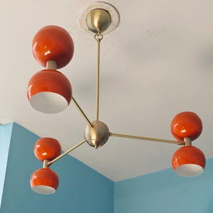 Puede incluir: Un candelabro de estilo retro con estructura de latón y cuatro pantallas globulares naranjas. Las pantallas tienen un acabado brillante e interiores blancos. La lámpara está montada en un techo blanco.