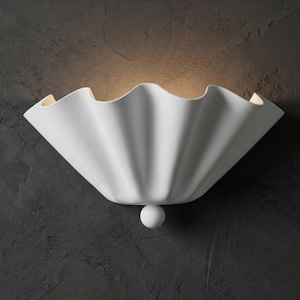 Może przedstawiać: Biały kinkiet ścienny o muszlowatym kształcie. Oprawa ma gładkie wykończenie i mały, kulisty detal na dole. Światło emituje ciepłą poświatę na ciemnoszarej ścianie.