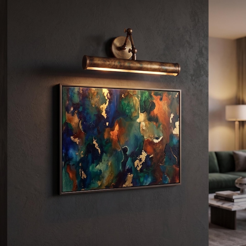 May include: An abstract painting with a gold frame, featuring a mix of blue, green, and orange hues, is illuminated by a bronze-toned picture light. The artwork is mounted on a dark gray wall, creating a sophisticated ambiance.