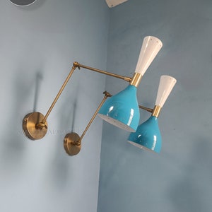 Puede incluir: Dos apliques de pared de latón con pantallas de esmalte azul y blanco. Las apliques tienen brazos ajustables y están montadas en una pared azul.