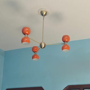 May include: A mid-century modern chandelier with five orange globe-shaped lights. The fixture has a brass finish and is mounted on a white ceiling. The lights are arranged on a starburst design.