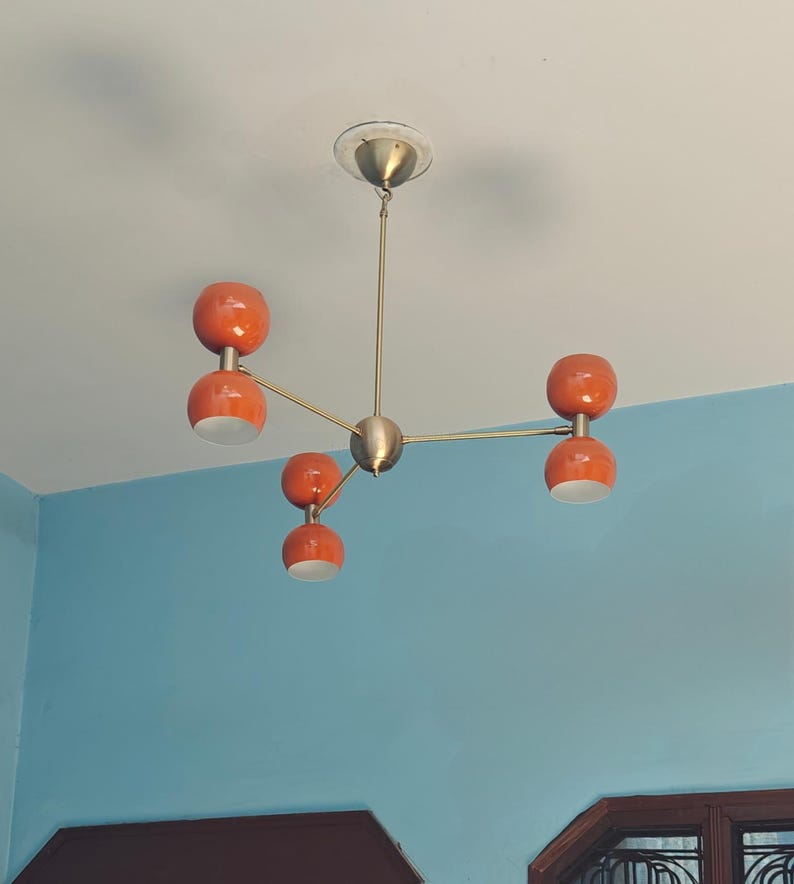 May include: A mid-century modern chandelier with five orange globe-shaped lights. The fixture has a brass finish and is mounted on a white ceiling. The lights are arranged on a starburst design.