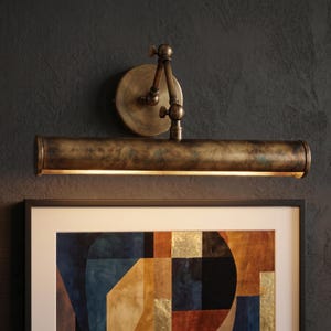 May include: A vintage-style picture light with an aged brass finish. The light has a long, cylindrical shade with a warm glow, mounted on an adjustable arm and a circular backplate. It is installed above a framed abstract painting.