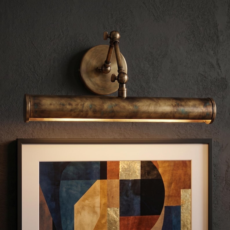 May include: A vintage-style picture light with an aged brass finish. The light has a long, cylindrical shade with a warm glow, mounted on an adjustable arm and a circular backplate. It is installed above a framed abstract painting.