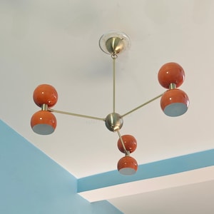 May include: A mid-century modern style chandelier with five orange globe shades and a brass finish. The light fixture is suspended from the ceiling and has a central sphere. The shades have a white interior.