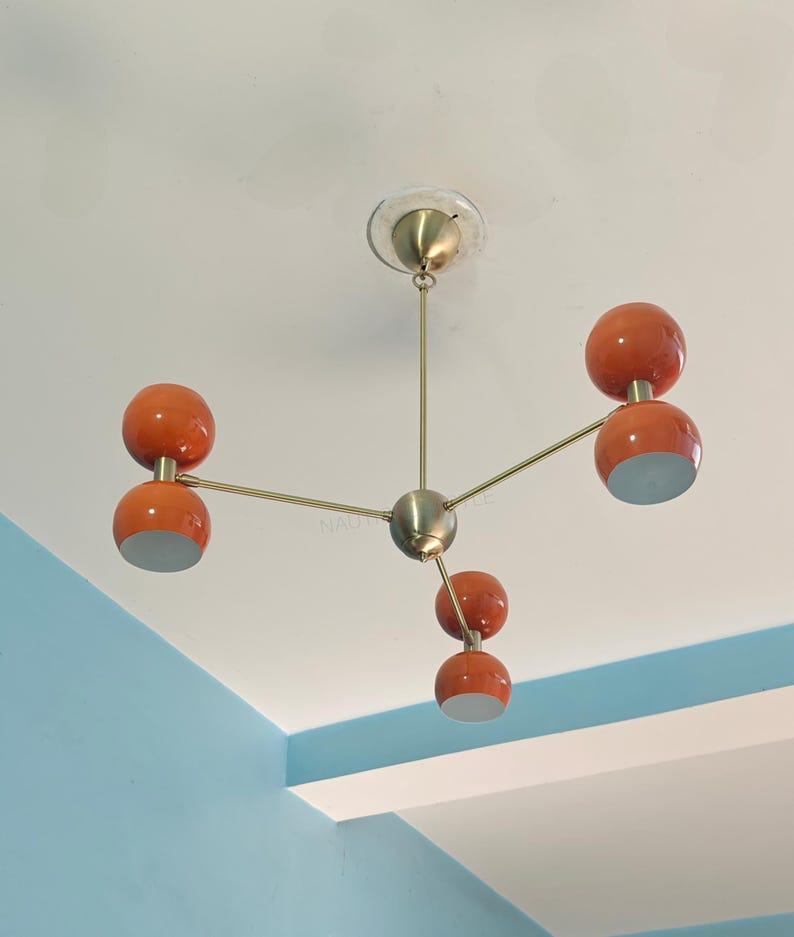 May include: A mid-century modern style chandelier with five orange globe shades and a brass finish. The light fixture is suspended from the ceiling and has a central sphere. The shades have a white interior.