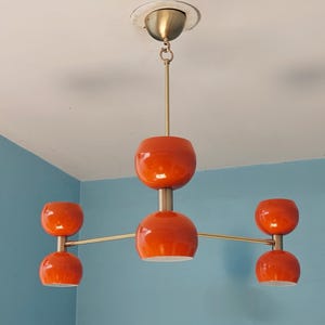 May include: A mid-century modern orange and brass chandelier with five spherical shades. The fixture is suspended from the ceiling by a brass rod and features a central sphere with four additional shades extending outwards.