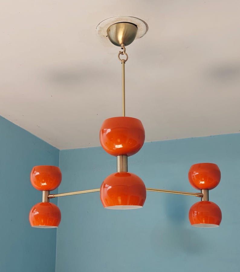 May include: A mid-century modern orange and brass chandelier with five spherical shades. The fixture is suspended from the ceiling by a brass rod and features a central sphere with four additional shades extending outwards.
