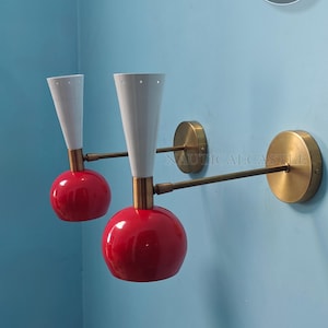May include: A pair of red and white wall sconces with brass accents. The sconces have a round red shade and a white cone-shaped shade. The sconces are mounted on a brass arm.