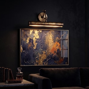 May include: An abstract painting with gold leaf accents and a brass picture light. The artwork features a textured surface with shades of blue and orange. The light fixture is mounted above the framed art, illuminating the piece.