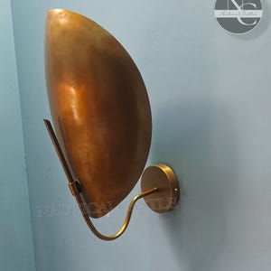 Puede incluir: Una lámpara de pared de latón con una pantalla curva y redondeada. La lámpara tiene un diseño simple y moderno.