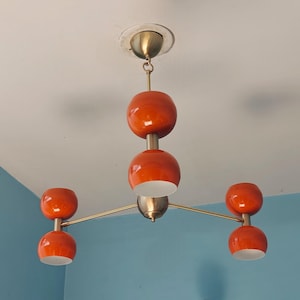 May include: A mid-century modern style chandelier with five orange globe-shaped lights. The fixture has a brass finish and is mounted on a white ceiling. The lights are arranged in a unique, eye-catching design.
