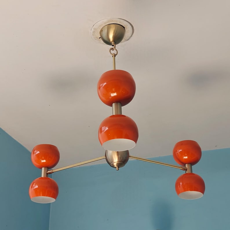 May include: A mid-century modern style chandelier with five orange globe-shaped lights. The fixture has a brass finish and is mounted on a white ceiling. The lights are arranged in a unique, eye-catching design.