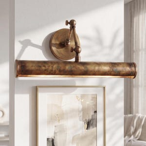 May include: A bronze-toned picture light fixture with an adjustable arm and a round backplate. The light has a long, cylindrical shade that emits a warm glow. The fixture is mounted on a white wall above a framed artwork.