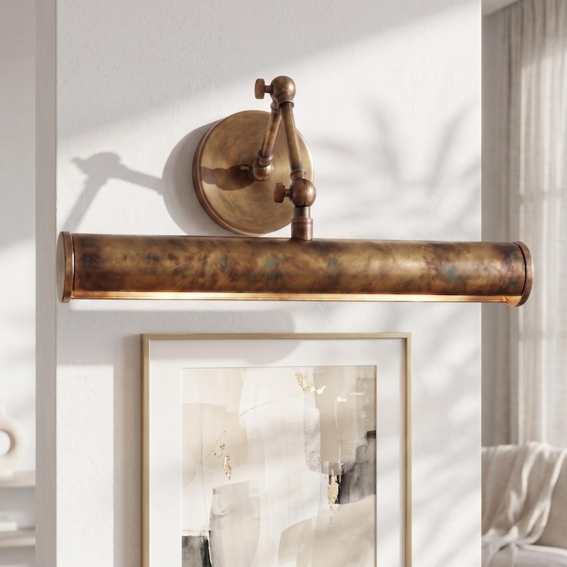May include: A bronze-toned picture light fixture with an adjustable arm and a round backplate. The light has a long, cylindrical shade that emits a warm glow. The fixture is mounted on a white wall above a framed artwork.