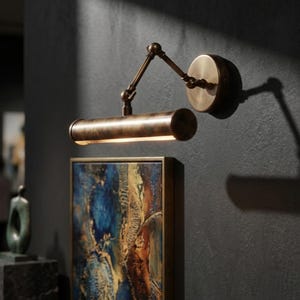 May include: A bronze-toned picture light with an adjustable arm and a circular wall mount. The light is positioned above a framed abstract painting with blue and brown hues, creating a warm, inviting ambiance. The fixture is designed for artwork illumination.