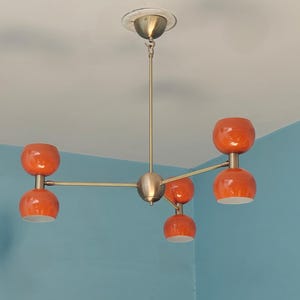 May include: A mid-century modern chandelier with five orange globe-shaped shades. The fixture has a brass finish and is mounted on a white ceiling. The orange shades have a glossy finish and are evenly spaced.