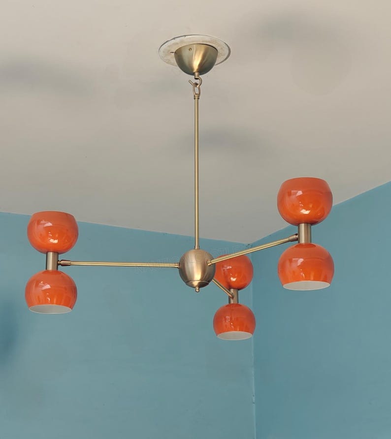 May include: A mid-century modern chandelier with five orange globe-shaped shades. The fixture has a brass finish and is mounted on a white ceiling. The orange shades have a glossy finish and are evenly spaced.