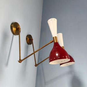 Peut inclure: Appliques murales de style moderne du milieu du siècle. Les appliques ont des abat-jours rouges en forme de cône avec des intérieurs blancs et de petites perforations. Les abat-jours sont associés à des sommets blancs et allongés. Les luminaires ont des bras et des supports muraux de couleur laiton.