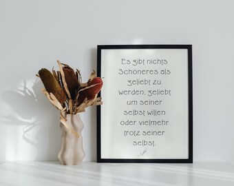 May include: A framed print with the German text "Es gibt nichts Schöneres als geliebt zu werden..." sits on a white surface. A beige vase with dried flowers is to the left. The frame is black.