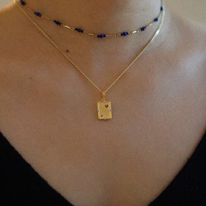 May include: A gold necklace with a small rectangular pendant featuring a red heart and small clear stones. The necklace is layered over a blue beaded chain.