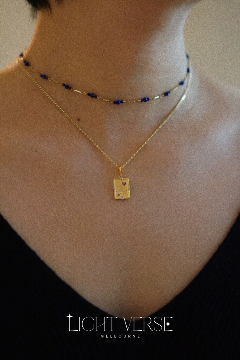 May include: A gold necklace with a small rectangular pendant featuring a red heart and small clear stones. The necklace is layered over a blue beaded chain.