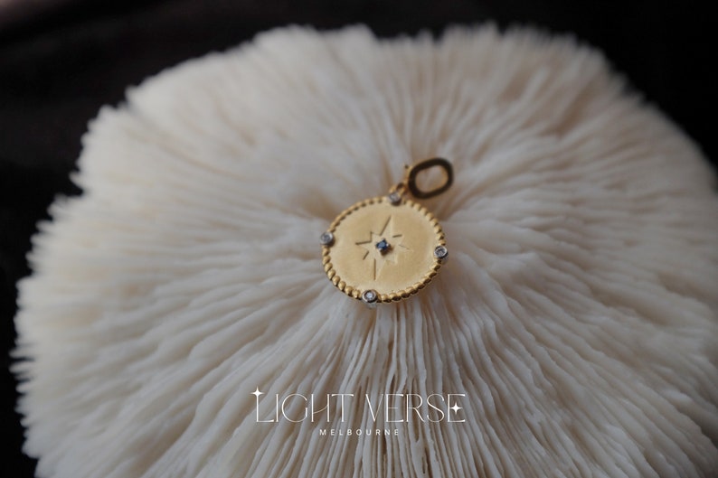 May include: Gold pendant necklace with a star design and small clear stones around the edge. The pendant is on a white coral-like surface.