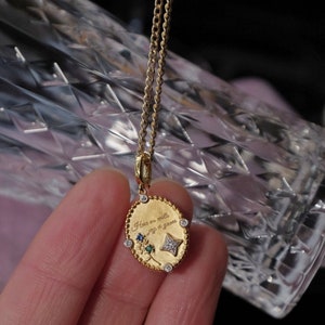 May include: A gold chain necklace with a small oval pendant. The pendant has a floral design with green, blue, and white gemstones. The pendant is engraved with the text "Here on earth, only a glance".