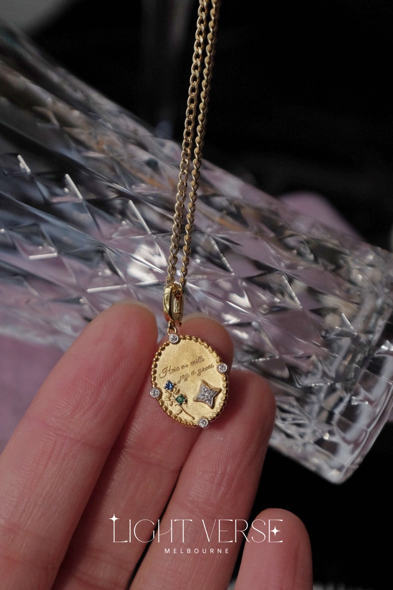 May include: A gold chain necklace with a small oval pendant. The pendant has a floral design with green, blue, and white gemstones. The pendant is engraved with the text "Here on earth, only a glance".
