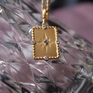 May include: Gold necklace with a rectangular pendant featuring a star design with a blue gemstone in the center. The pendant is surrounded by small clear gemstones.
