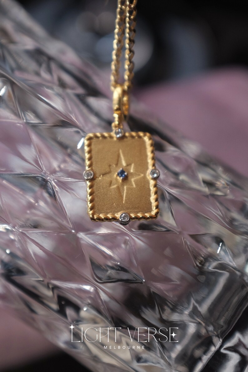 May include: Gold necklace with a rectangular pendant featuring a star design with a blue gemstone in the center. The pendant is surrounded by small clear gemstones.
