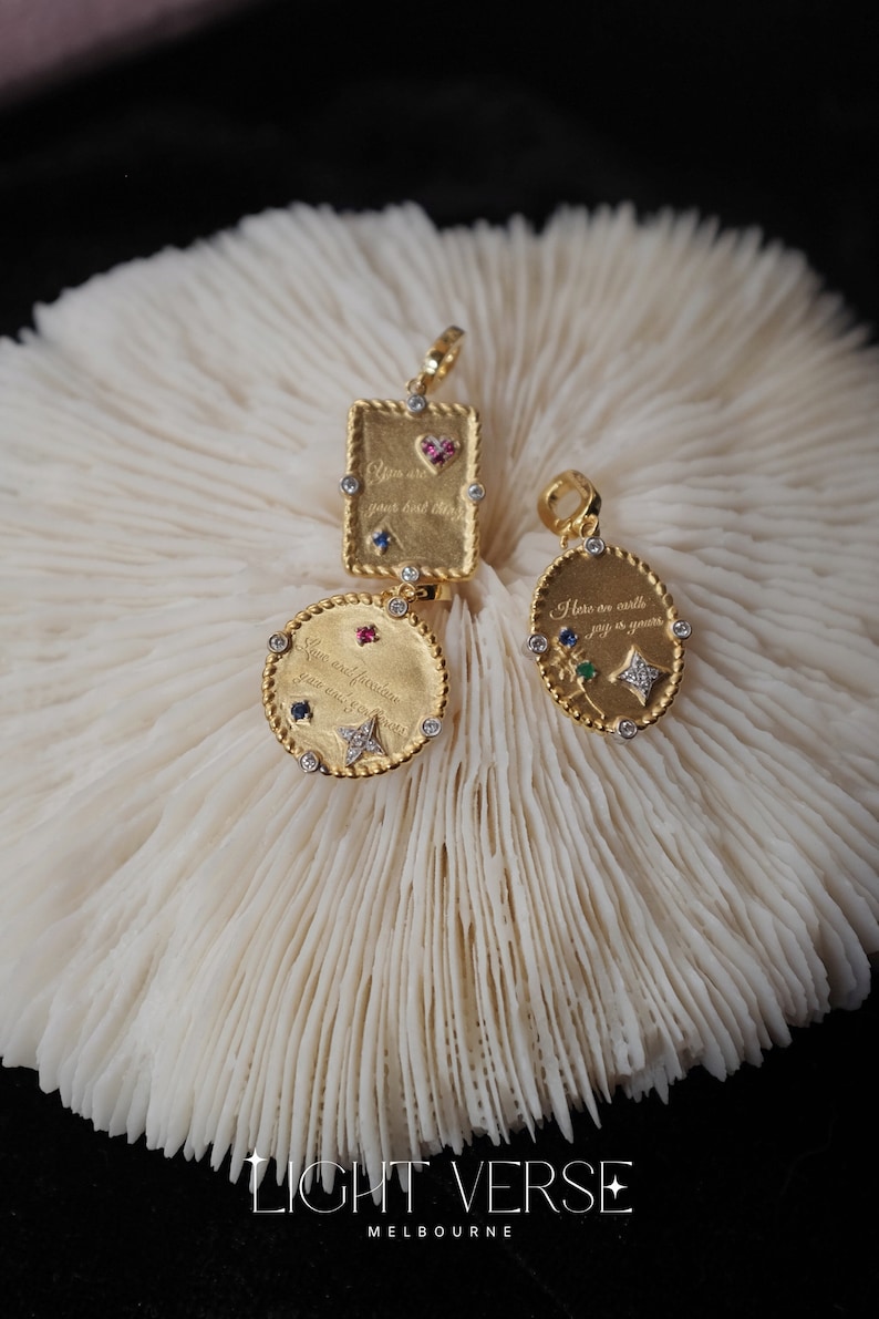 May include: Three gold pendants with colorful gemstones and engraved messages. The pendants are on a white coral background. The messages on the pendants are "You are your best friend", "Here are stars, joy is years", and "Silver and freedom you and forever".