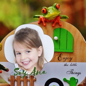 May include: A wooden picture frame with a photo of a smiling girl. The frame has a green door, a frog figurine, and the words "Enjoy the little Things". The name "Soy Ana" is also visible.