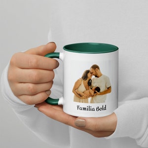 Könnte beinhalten: Eine weiße Keramiktasse mit grünem Rand und Henkel. Die Tasse zeigt ein Foto einer dreiköpfigen Familie, ein Paar hält ein Baby, und den Text "Familia Bold".