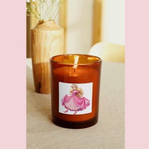 May include: A brown glass candle with a white label featuring a cartoon princess in a pink dress. The candle is lit and the flame is visible.