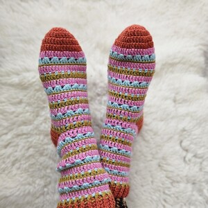 Op de afbeelding: Een paar gehaakte sokken met een gestreept patroon in tinten roze, blauw, bruin en rood. De sokken worden gedragen door iemand met luipaardprint leggings.
