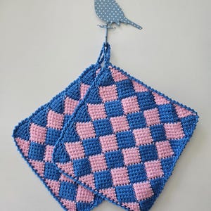 Puede incluir: Dos salvamanteles cuadrados con un patrón de tablero de ajedrez en azul y rosa. Cuelgan de un gancho de pared azul con forma de pájaro. Los salvamanteles son de material tejido.