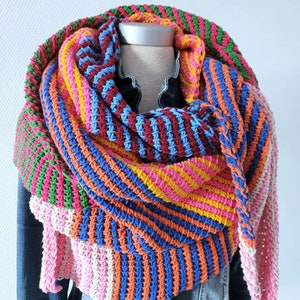 Op de afbeelding: Een kleurrijke gestreepte gehaakte sjaal in roze, oranje, blauw, geel en groen. De sjaal is gevouwen en gedrapeerd over een mannequin met een spijkerjack.