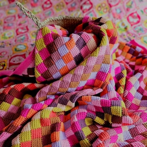 Puede incluir: Una manta de crochet colorida con un patrón de cuadros en tonos de rosa, morado, naranja, marrón y verde. La manta está doblada y drapeada sobre una cesta de mimbre.