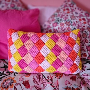 Puede incluir: Una almohada de crochet colorida con un patrón de diamantes en tonos de rosa, amarillo y naranja. La almohada está sentada en una cama con una colcha estampada con flores.