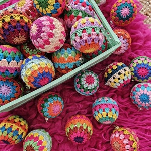 May include: A collection of colorful crocheted ornaments in a green crate and scattered on a pink fuzzy surface. The ornaments are made with different colored yarn and have a variety of patterns.
