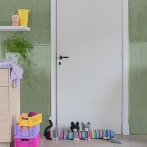 Op de afbeelding: Een kleurrijke gehaakte kattendeurstop met drie gehaakte katten, een zwarte, een grijze en een witte, liggend op een grijze vloer voor een witte deur.