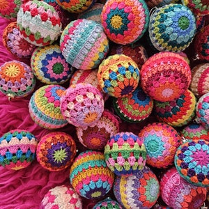 Op de afbeelding: Een verzameling kleurrijke gehaakte ornamenten in verschillende patronen en kleuren, waaronder rood, groen, blauw, geel en roze. De ornamenten zijn willekeurig gerangschikt op een roze pluche oppervlak.